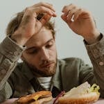 A bearded man in casual attire is looking at a deconstructed hamburger on a table.